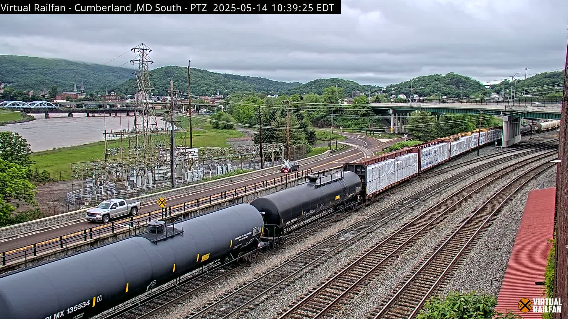 A long freight train carrying tanker cars and cargo passes under a bridge on a cloudy day near a river and a town nestled in a valley.