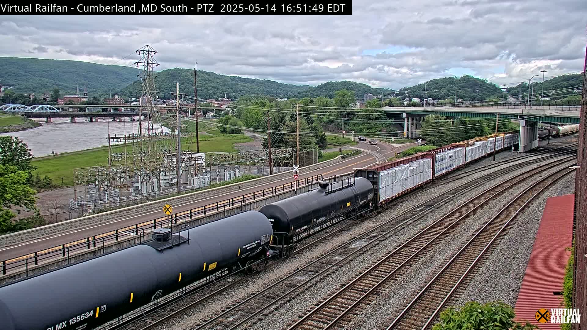 A freight train carrying tanker cars and boxcars is moving along railroad tracks under an overcast sky, with a river, bridge, and town visible in the background.