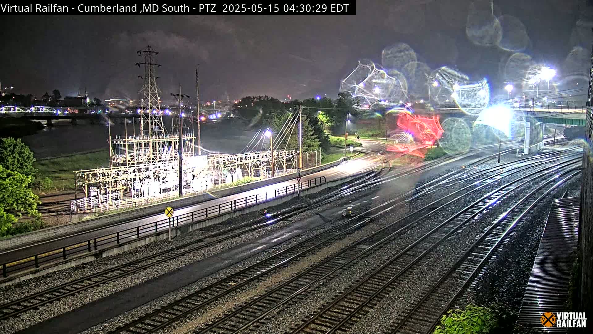 A nighttime view of rain-slicked train tracks near a power substation and a river, under a dark, cloudy sky.