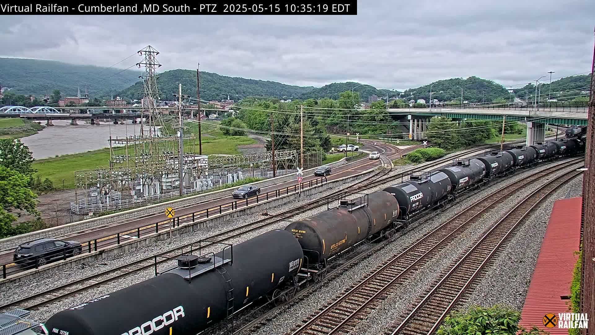 A long freight train of tanker cars moves along railroad tracks beside a river and road under an overcast sky, with a bridge and power station visible in the background.