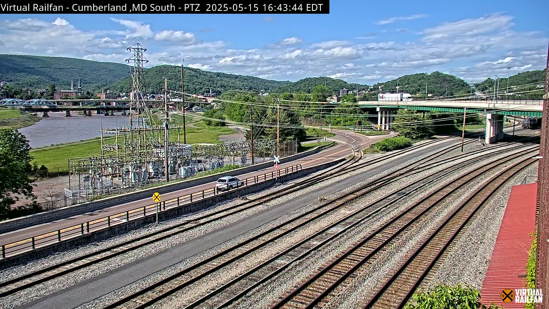 A sunny day shows a view of railroad tracks, a road, and an electrical substation beside a river, with a town and hills in the background.