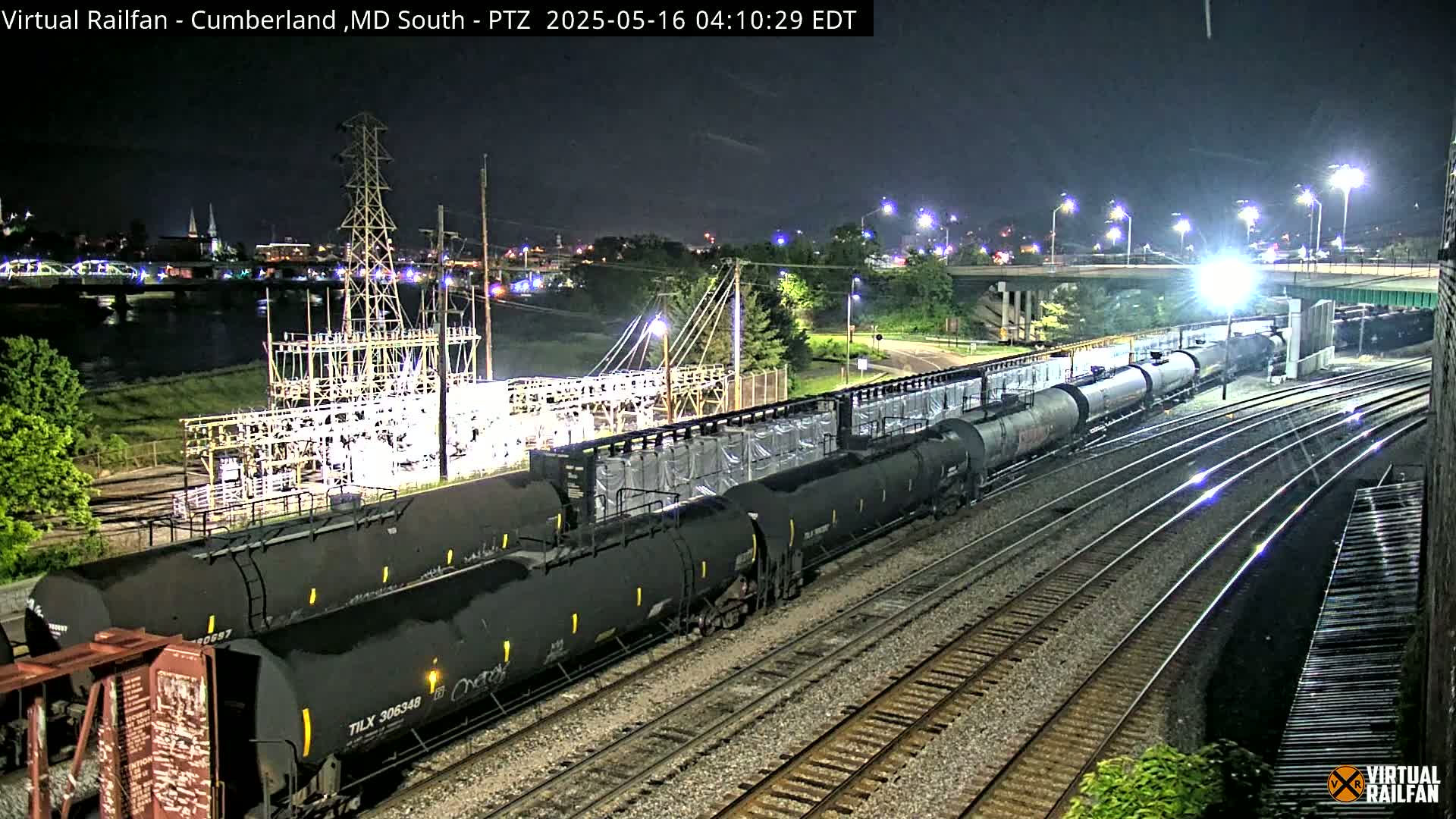 At night, a long freight train of tanker cars is seen parked on railroad tracks next to a power substation and a river, with city lights in the background.