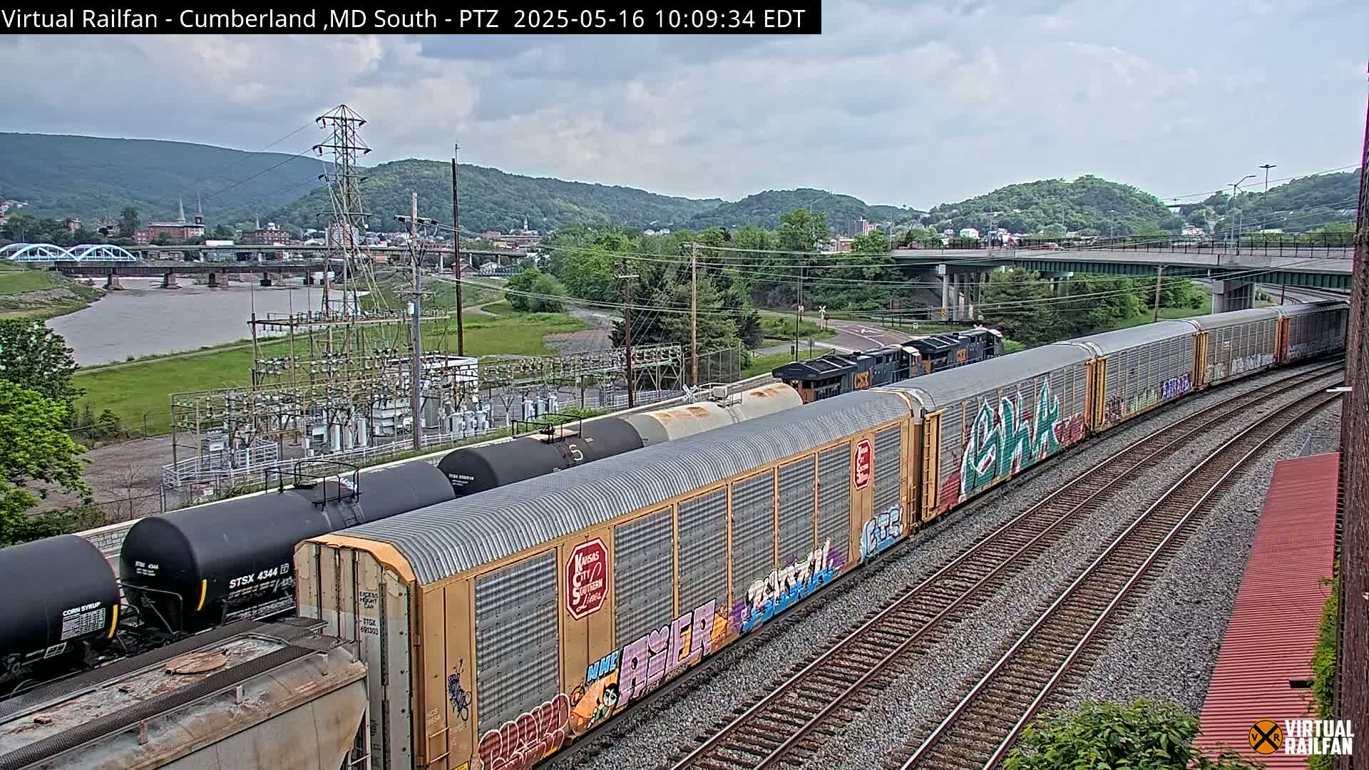 A long freight train with graffiti-covered cars travels along railroad tracks beside a river, under an overcast sky, past an industrial area and a bridge.