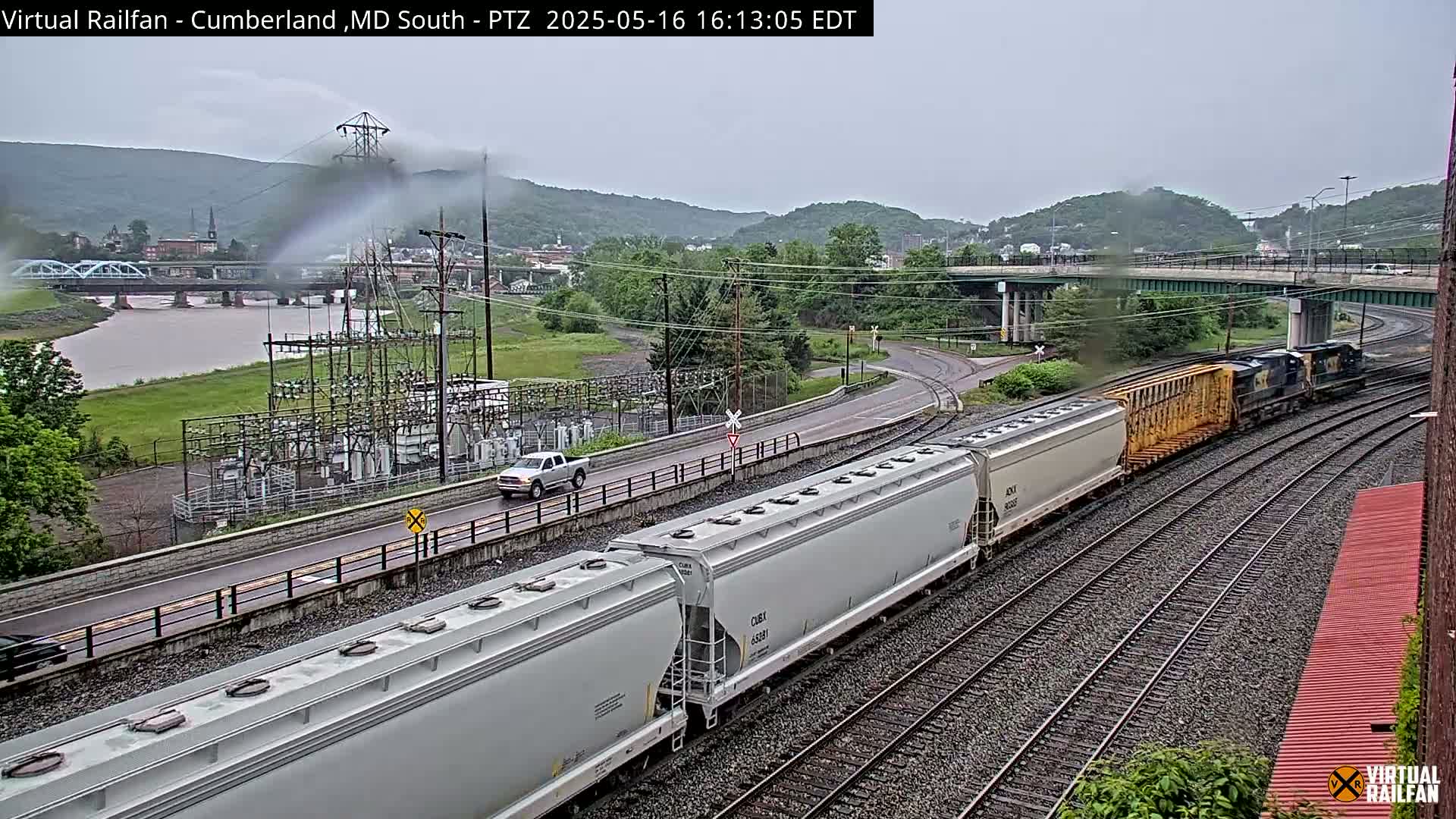 A long freight train passes under an overpass on a cloudy day, alongside a river and a road with vehicles.