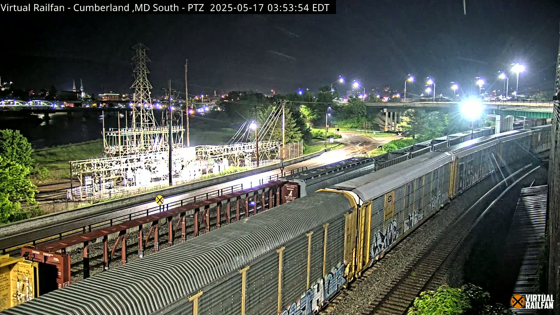 A long freight train is passing through a rail yard at night near a river and under a highway overpass, with a power substation visible in the background.