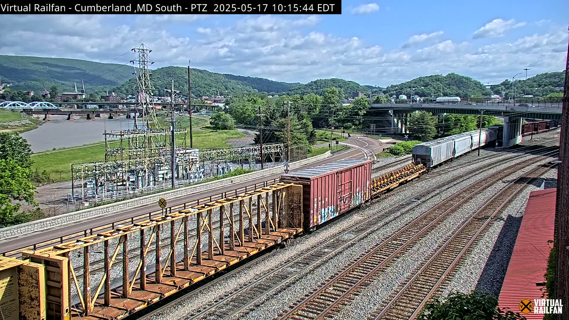 A freight train with various cars is moving along multiple parallel train tracks under a partly cloudy sky, passing near a river, power station, and bridge in a valley.