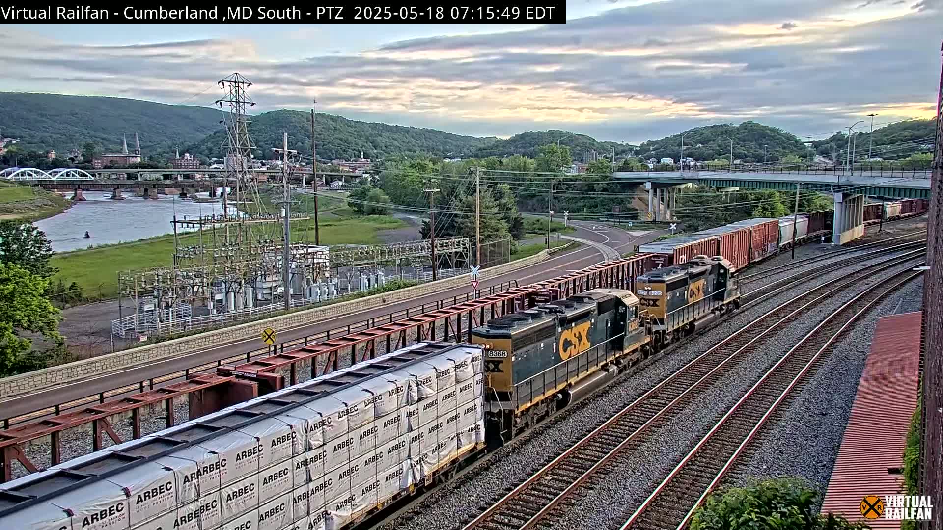 A freight train carrying lumber passes under a bridge near a river and power station under partly cloudy skies.