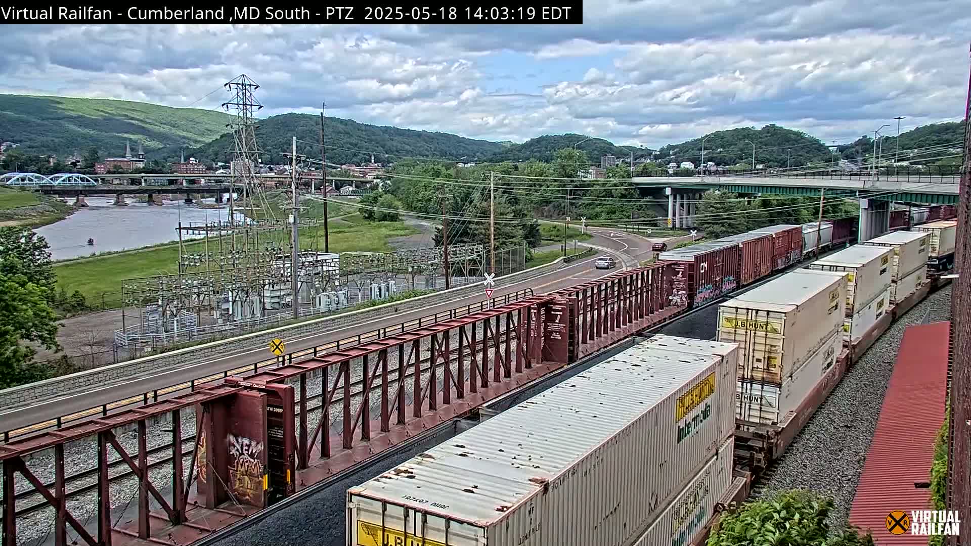 A long freight train carrying shipping containers is seen next to a river and highway bridge under a partly cloudy sky.