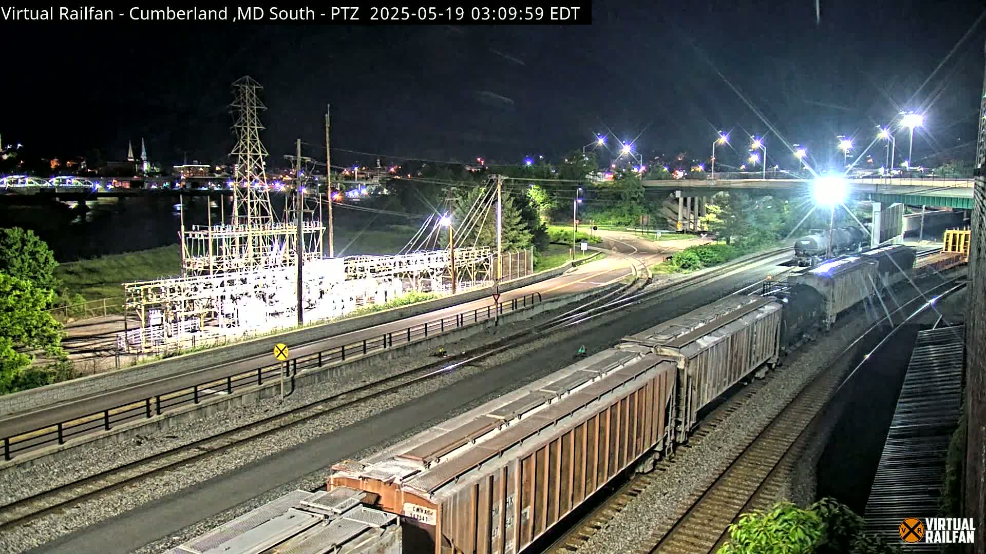 At night, a long freight train is visible on a set of train tracks near a power substation and a highway overpass, under a clear sky.