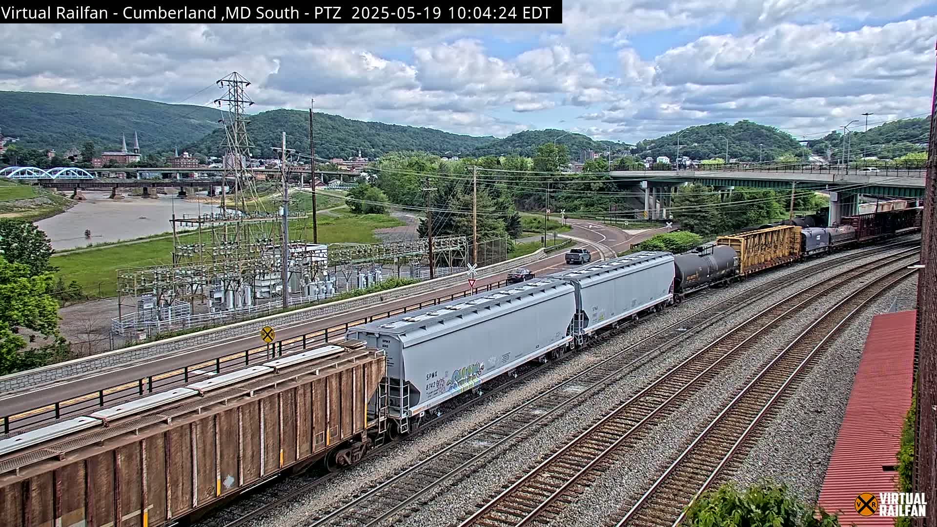 A long freight train passes through a valley on a partly cloudy day, near a river, a bridge, and a power substation.
