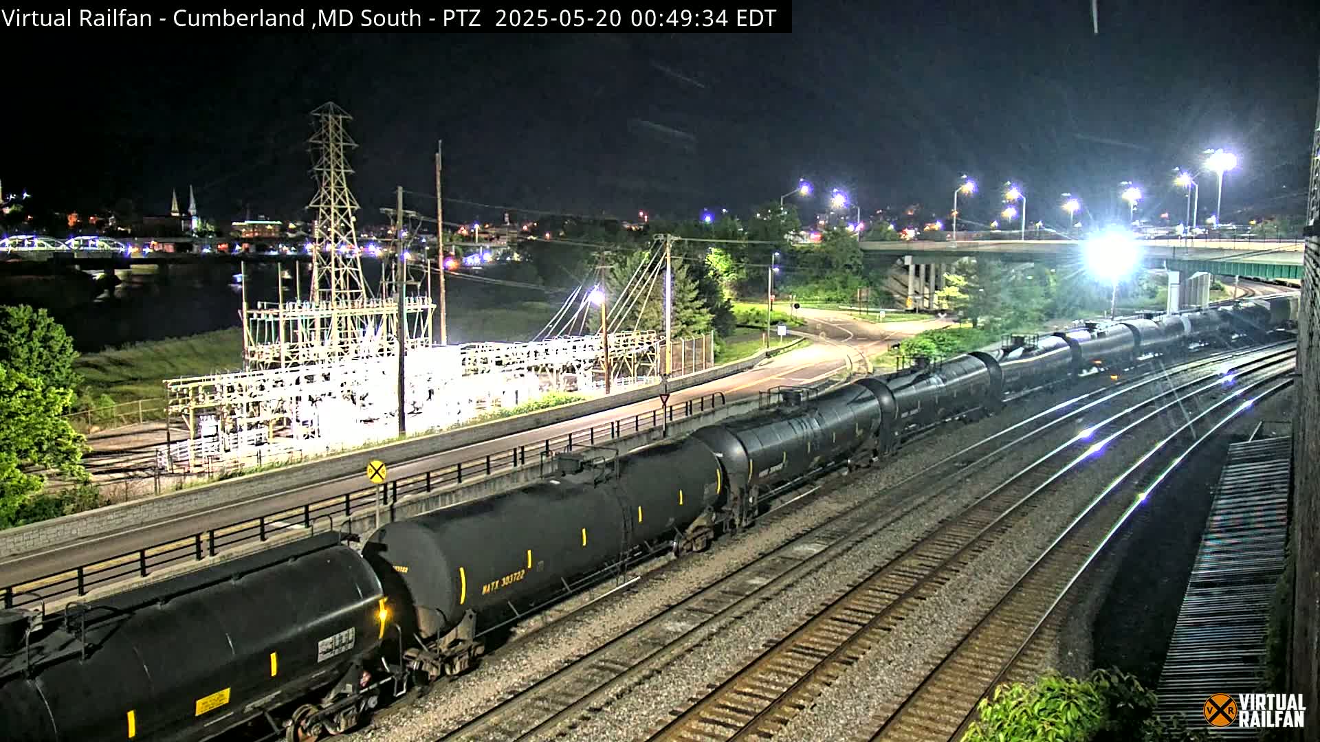 At night, a long train of dark tanker cars moves along multiple railroad tracks near a power station and a river, under well-lit conditions.