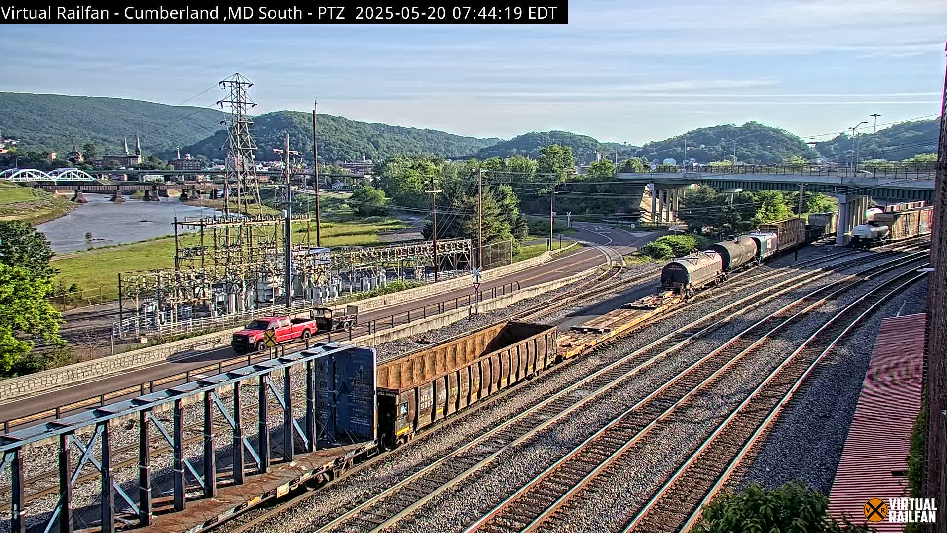 A partly sunny day reveals a view of railroad tracks with several train cars, a red pickup truck on a road next to a power substation, and a river and bridge in the background, with hills beyond.