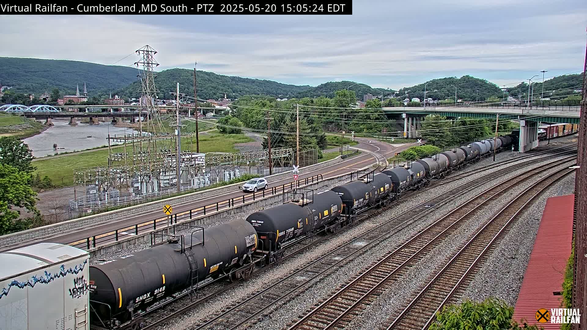 A long train of tanker cars is traveling along a set of railway tracks next to a river, under an overcast sky.