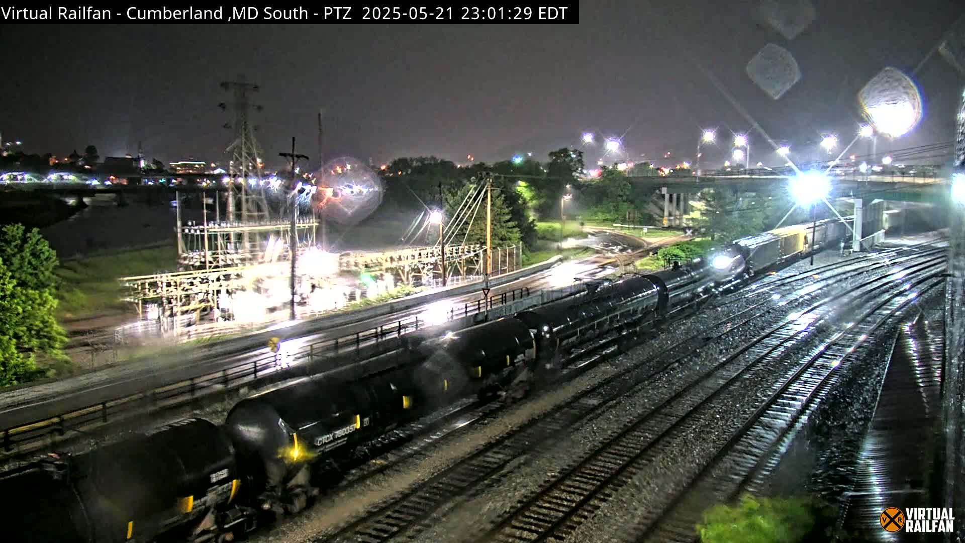 A nighttime, rainy view of a train of tanker cars on railroad tracks near an electrical substation and a roadway under a bridge.
