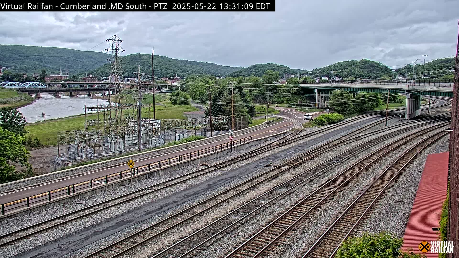 A high-angle view of a river, a bridge, railroad tracks, and a power substation under an overcast sky.