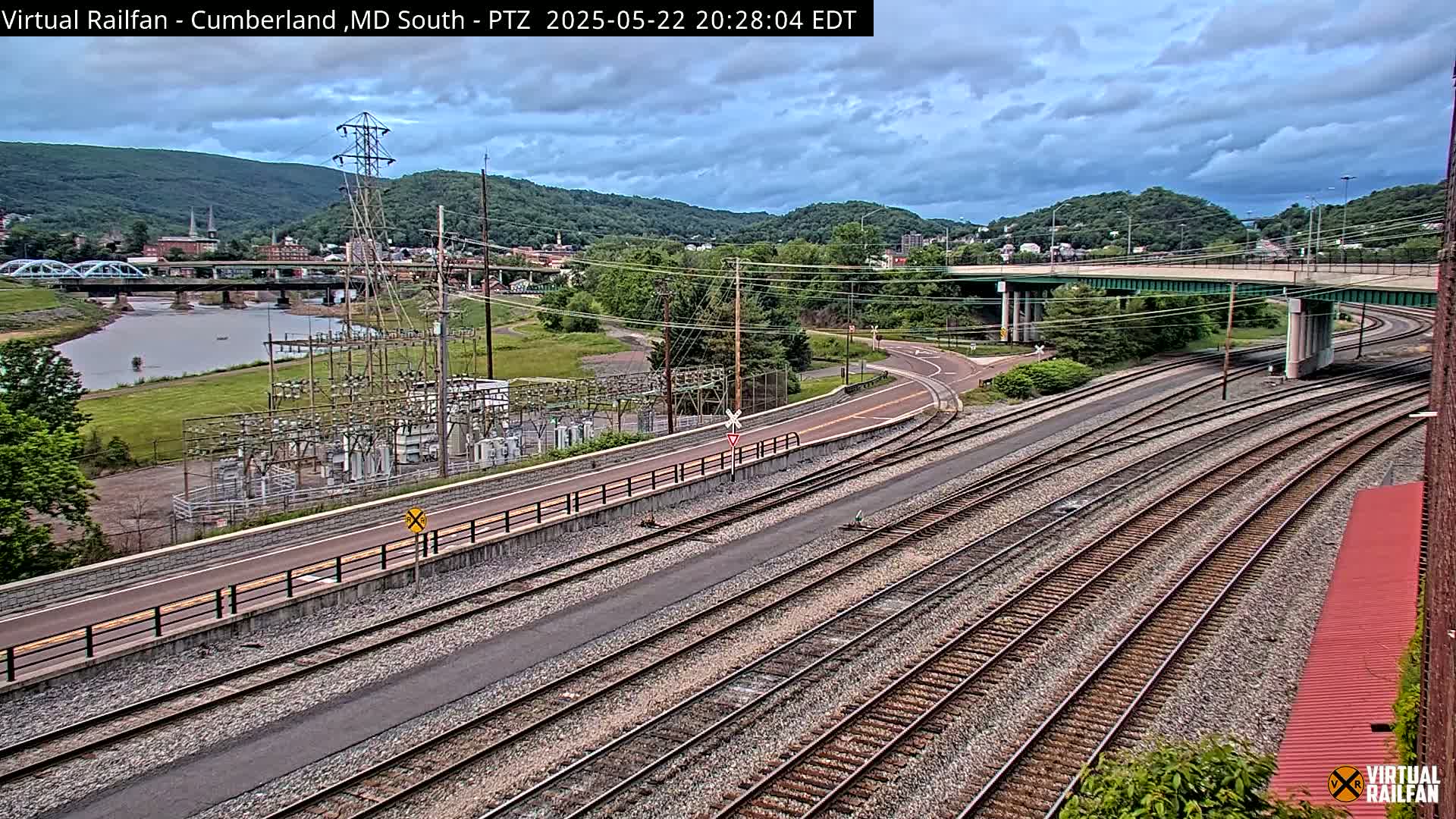 A high-angle view shows a river, a bridge, railroad tracks, a road, and an electrical substation under a partly cloudy sky.