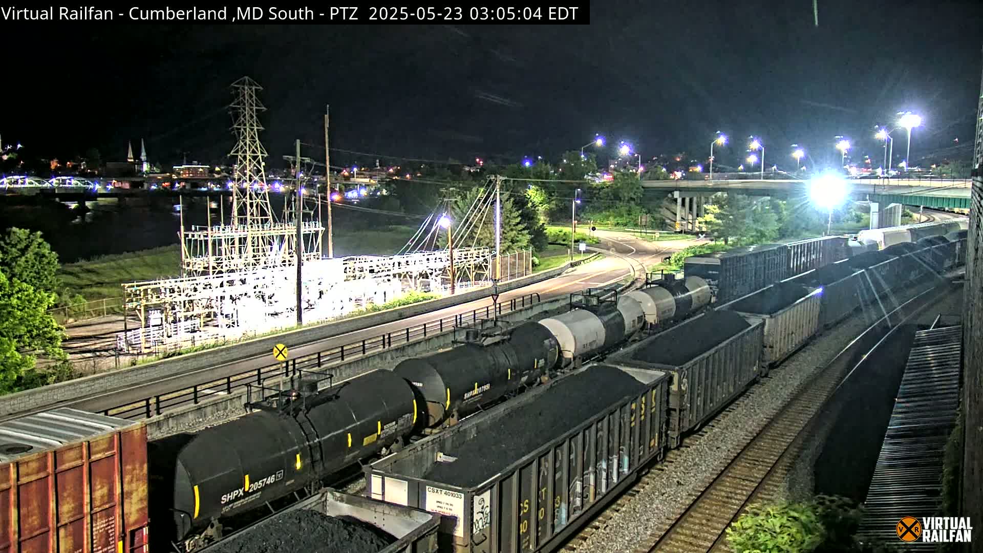 At night, a long freight train carrying coal and other materials is seen passing an electrical substation near a river and highway overpass.