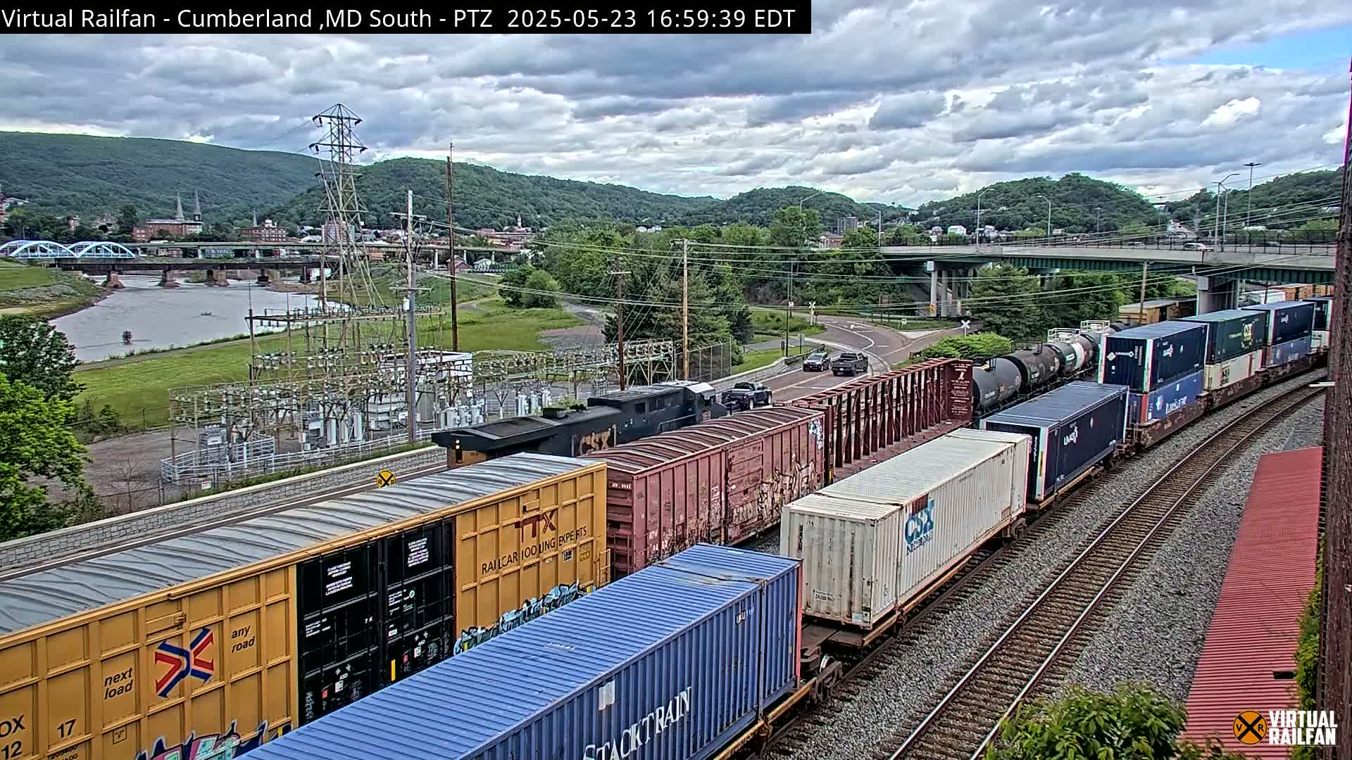 A long freight train with various containers and tanker cars is traveling along railroad tracks under partly cloudy skies, with a river, bridge, and town visible in the background.