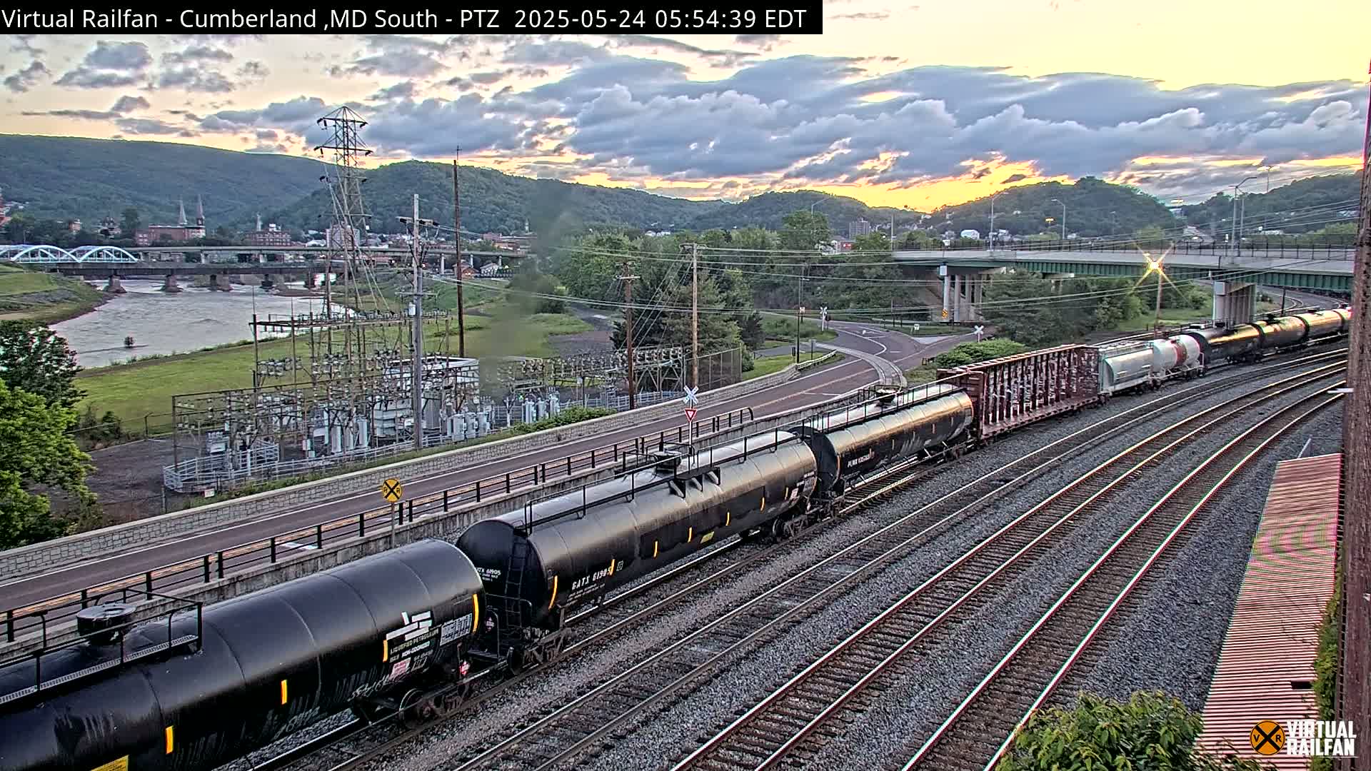 A long freight train is traveling along a set of railroad tracks under a partly cloudy sunset sky, with a river and a bridge visible in the background.