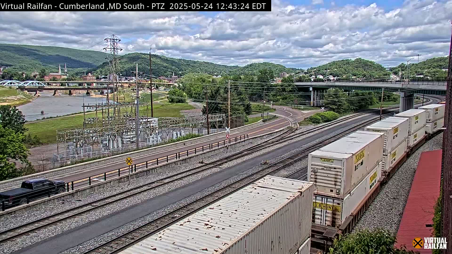 A partly cloudy day reveals a train carrying several white shipping containers on tracks next to a road and river, with a power station and a bridge in the background.