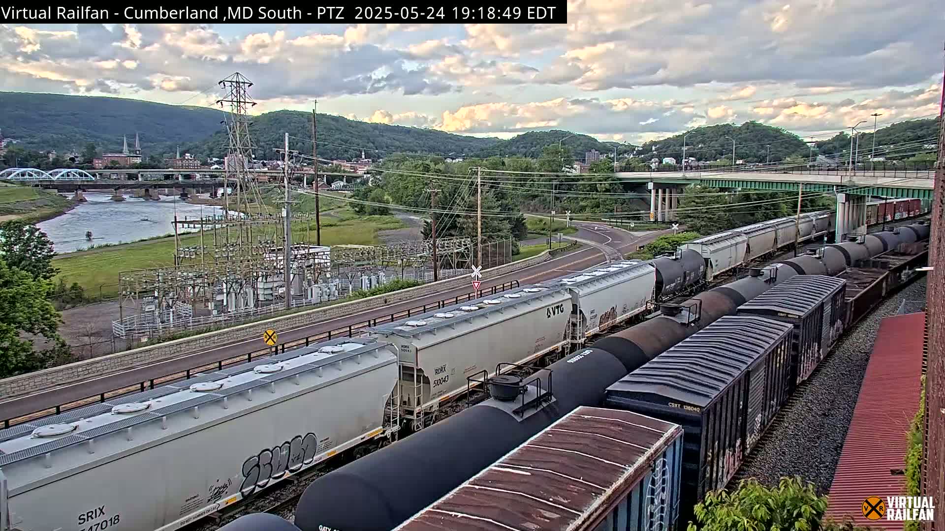 A long freight train, consisting of various boxcars and tanker cars, is traveling along railroad tracks under a partly cloudy evening sky, with a river and town visible in the background.
