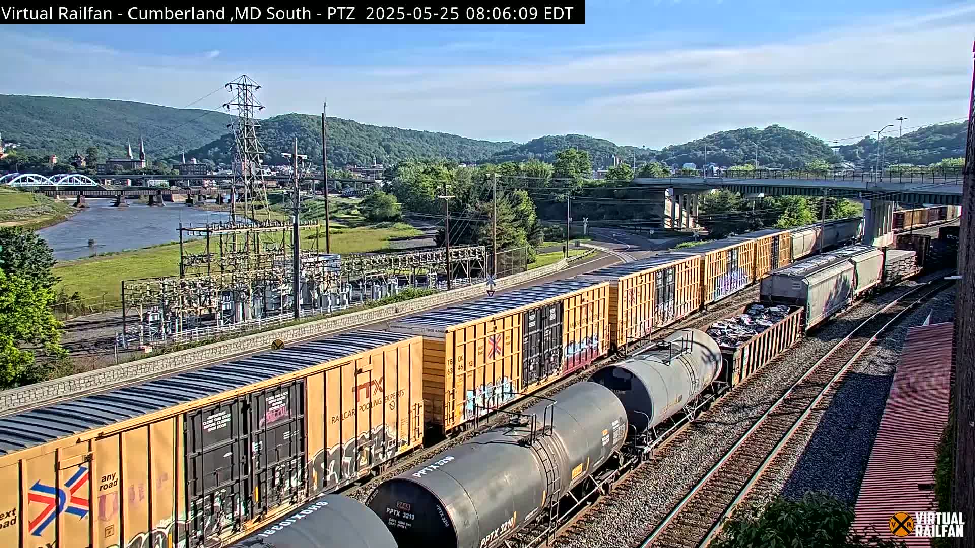 A long freight train carrying various cargo is parked on train tracks near a river and power station, under a mostly clear sky.
