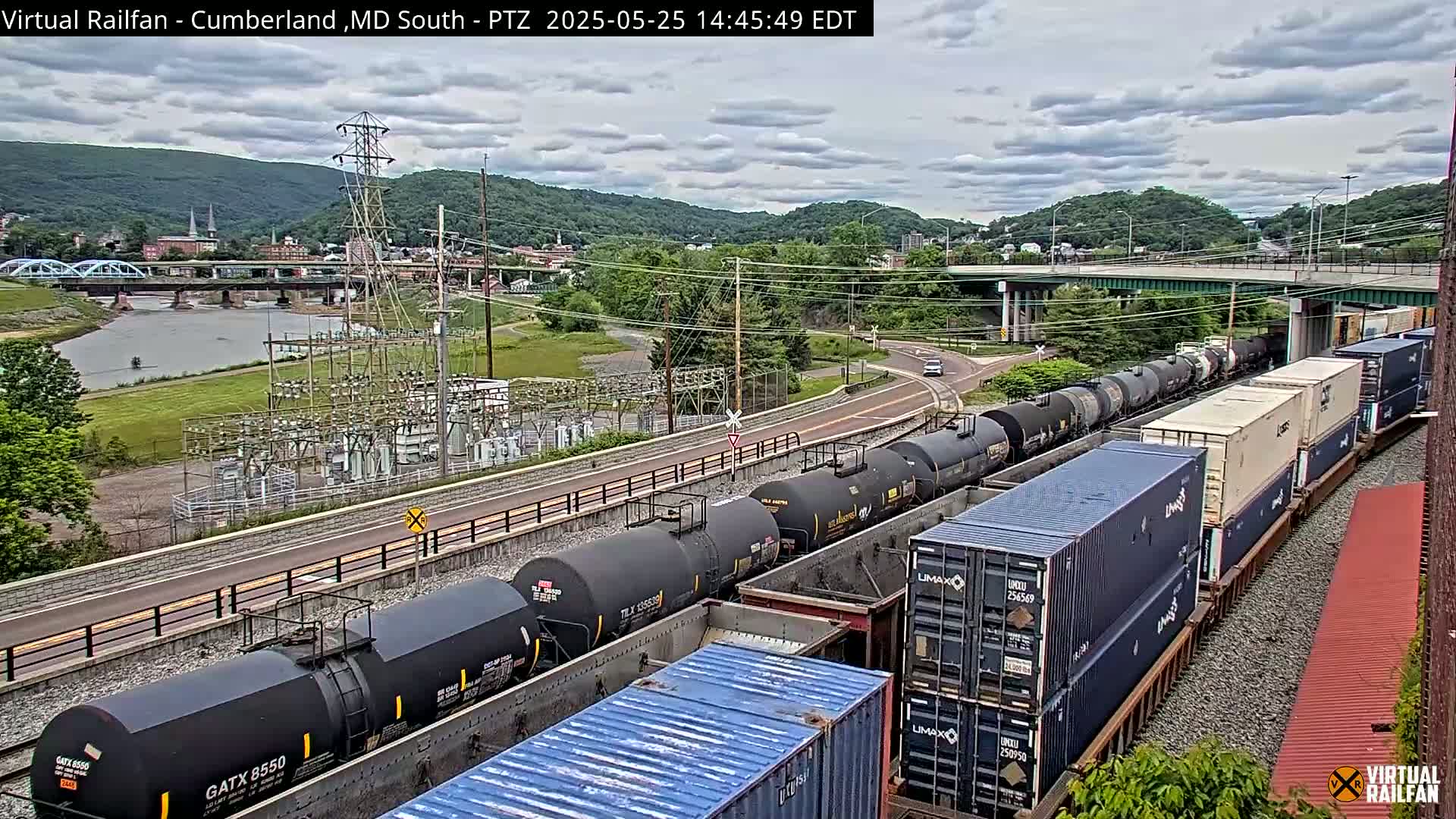 A long freight train carrying tanker cars and shipping containers is parked near a river, under a partly cloudy sky, with a bridge and power station in the background.