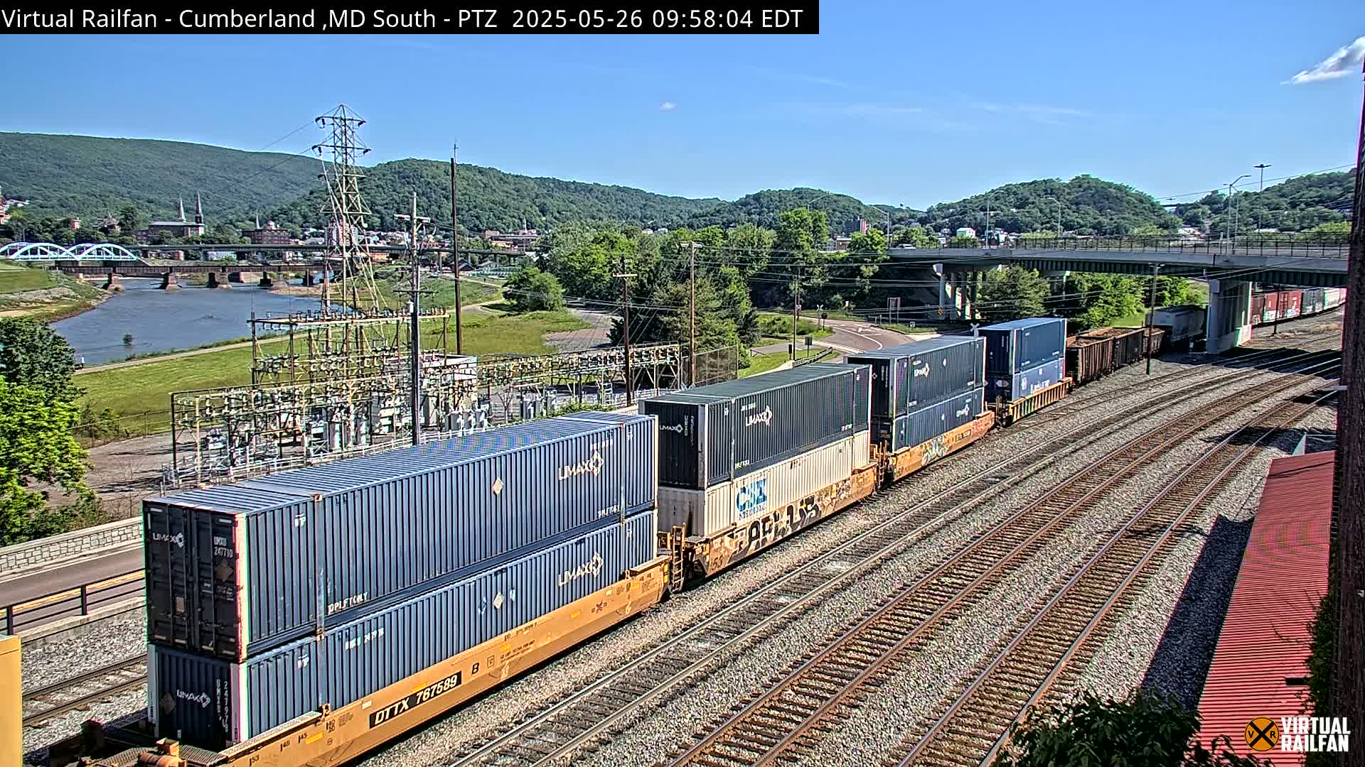 A freight train carrying stacked shipping containers moves along railroad tracks under a sunny sky, with a river, bridges, and hills in the background.