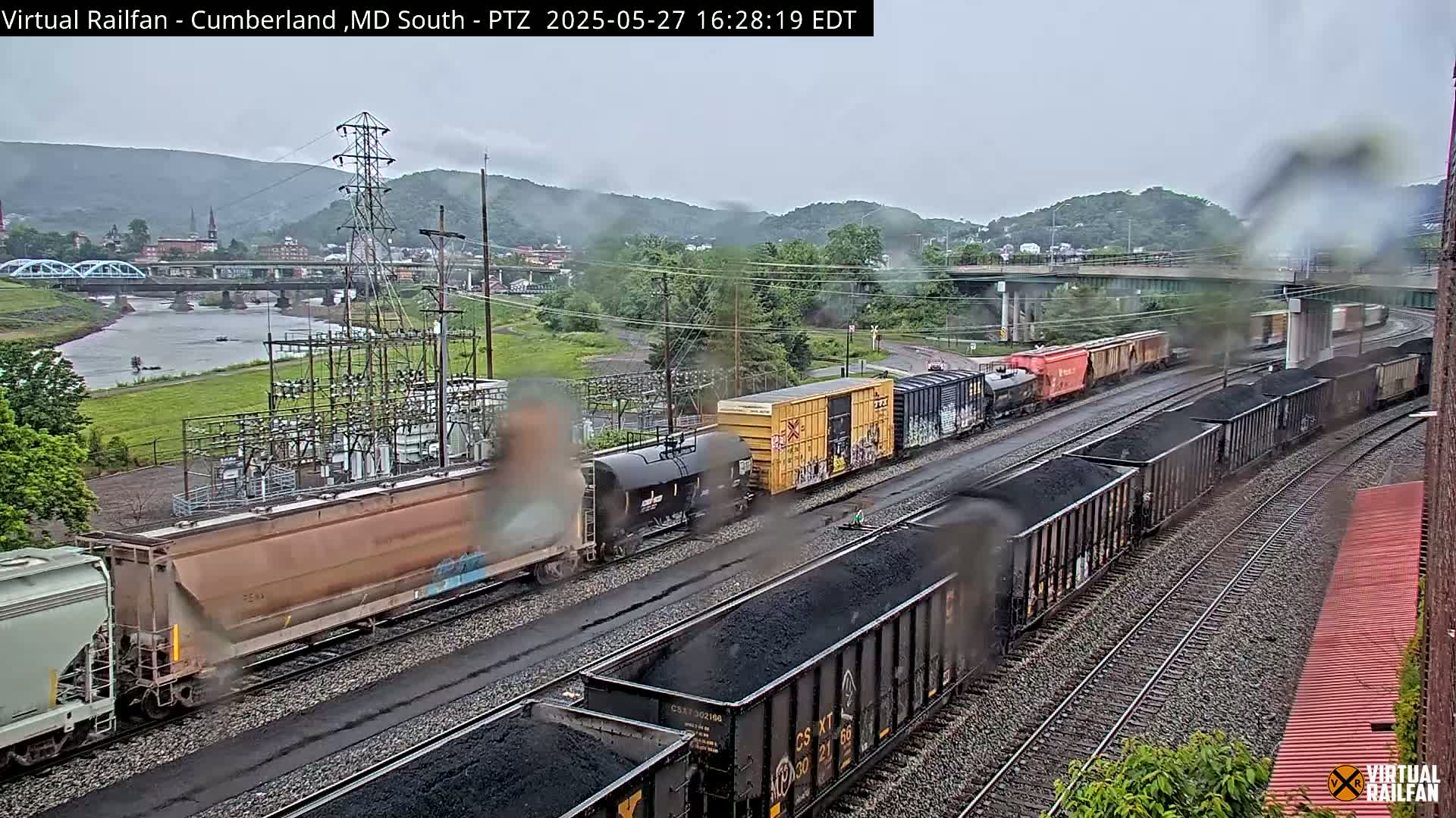 A long freight train carrying coal is seen on a railway track under an overcast sky, with a river and a town visible in the background.