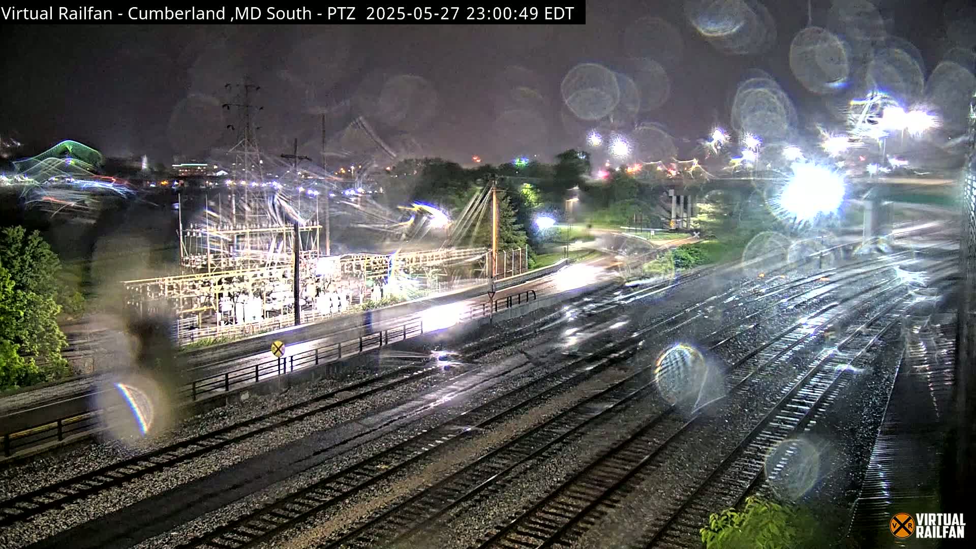 A nighttime view of rain-slicked railroad tracks next to a roadway and electrical substation, with blurry lights and raindrops on the camera lens.