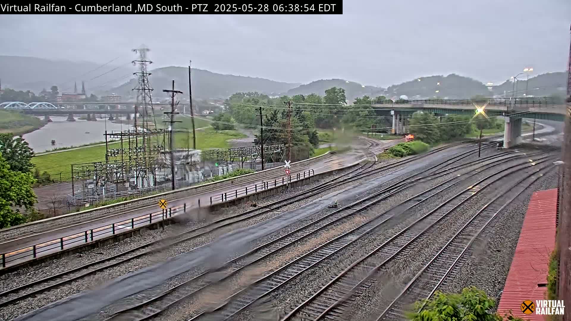 A rainy view of multiple train tracks intersecting near a river and a bridge, with a power substation and green hills in the background.
