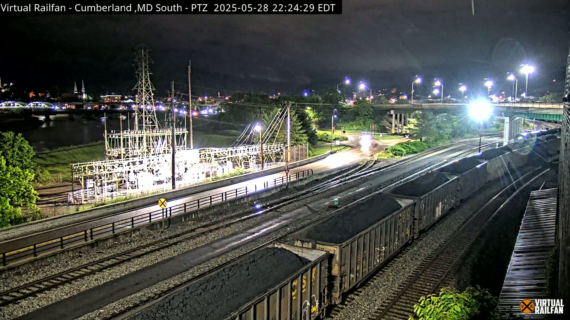 A nighttime view of a railway yard with several coal cars, an electrical substation, and a river in the background under a partly cloudy sky.