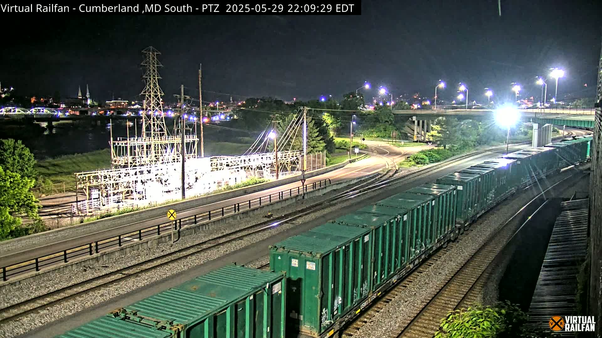 A long freight train with teal-colored containers moves along railroad tracks at night, passing near a power substation and a river with a city skyline in the distance.