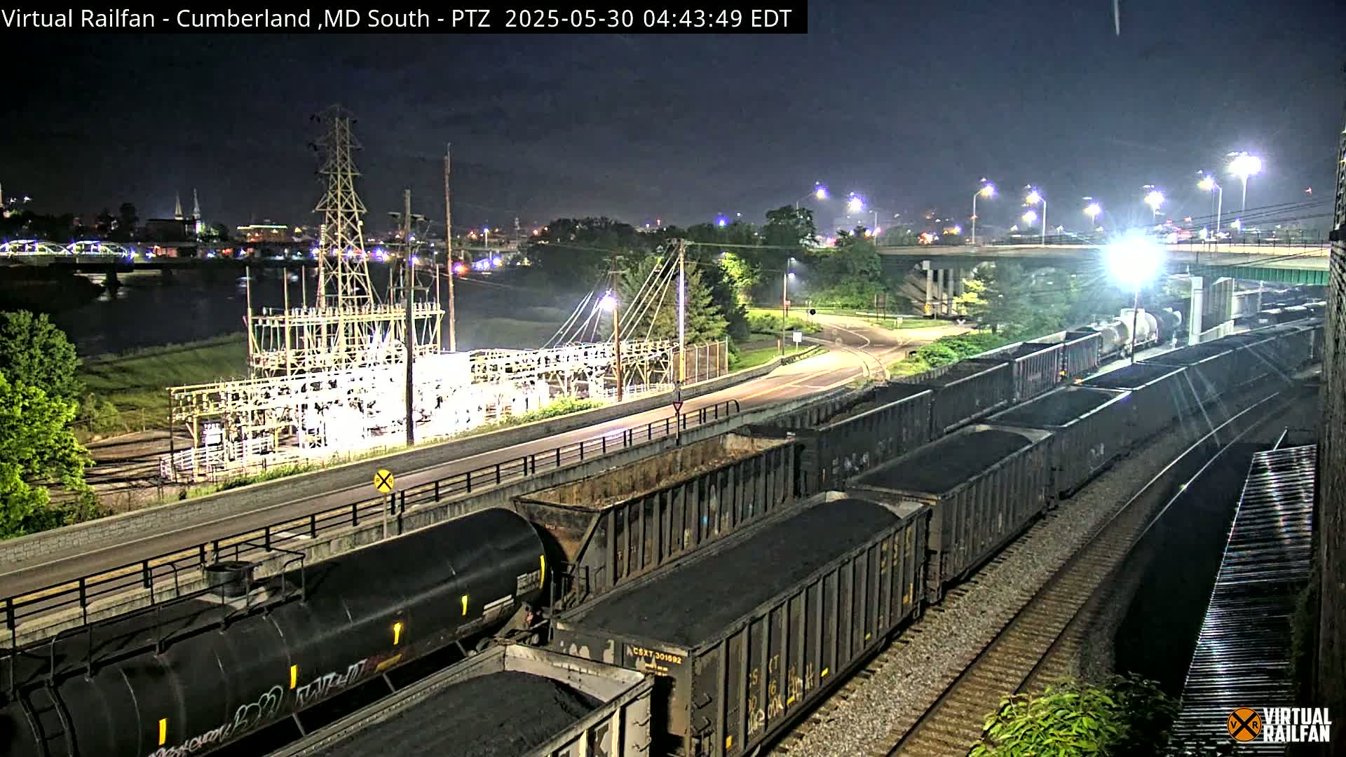At night, a long train carrying dark cargo is parked beside a roadway next to a river, under illuminated streetlights, with an electrical substation nearby.