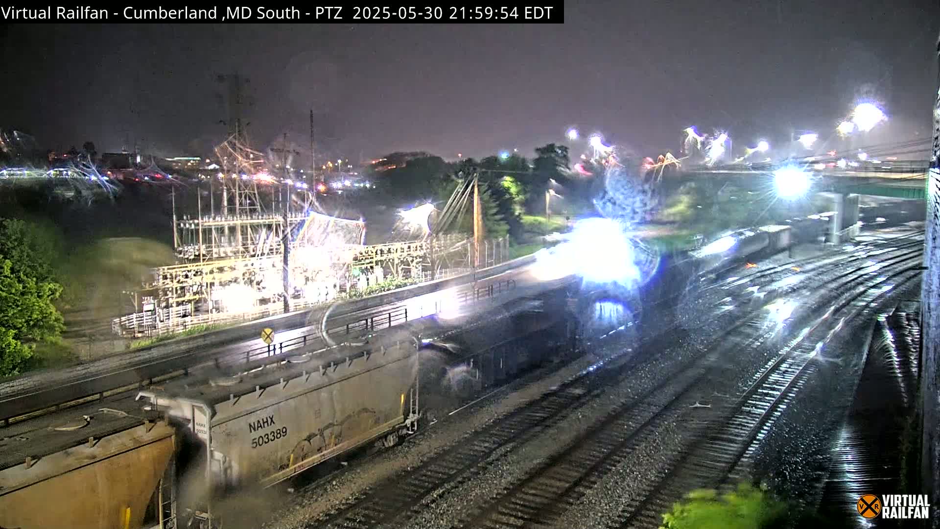 A nighttime view of a rain-soaked train yard with two freight trains, one partially obscured by rain and light, passing by an electrical substation under a dark, stormy sky.