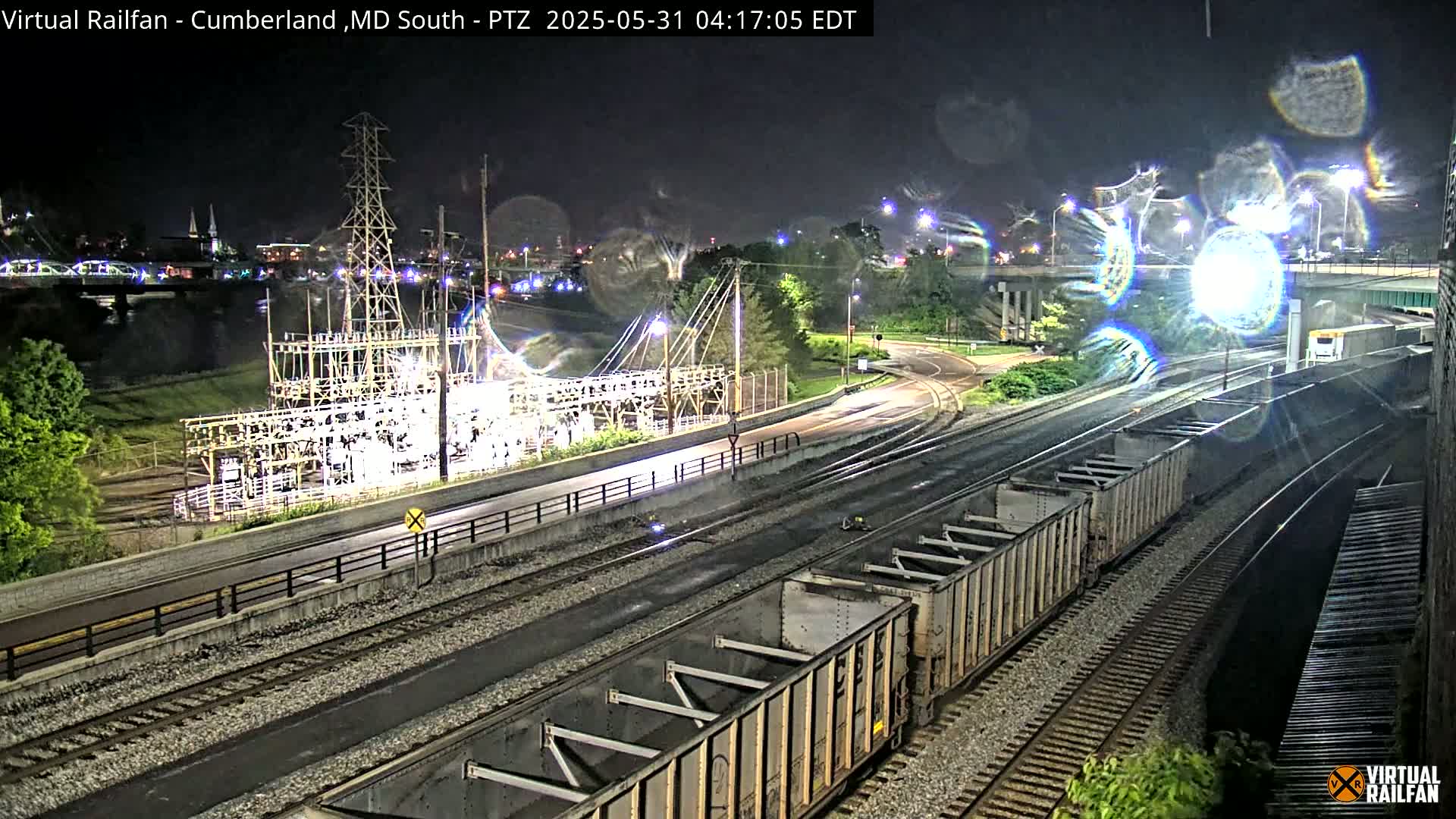 At night, a train carrying several boxcars sits on a set of railroad tracks next to a highway, near an electrical substation and a river, under a partly cloudy sky.
