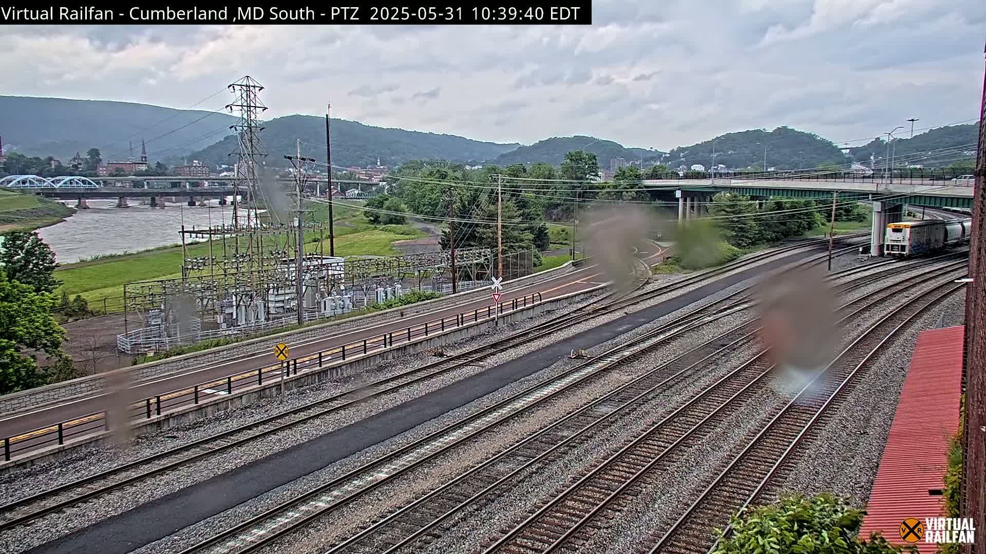 A train passes by a river and a power substation under an overcast sky.