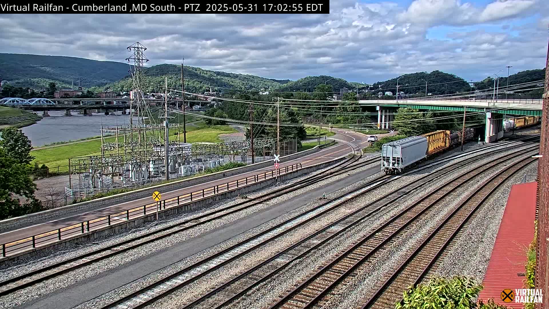 A freight train passes under a bridge near a river and power station, on a partly cloudy day with mountains in the background.