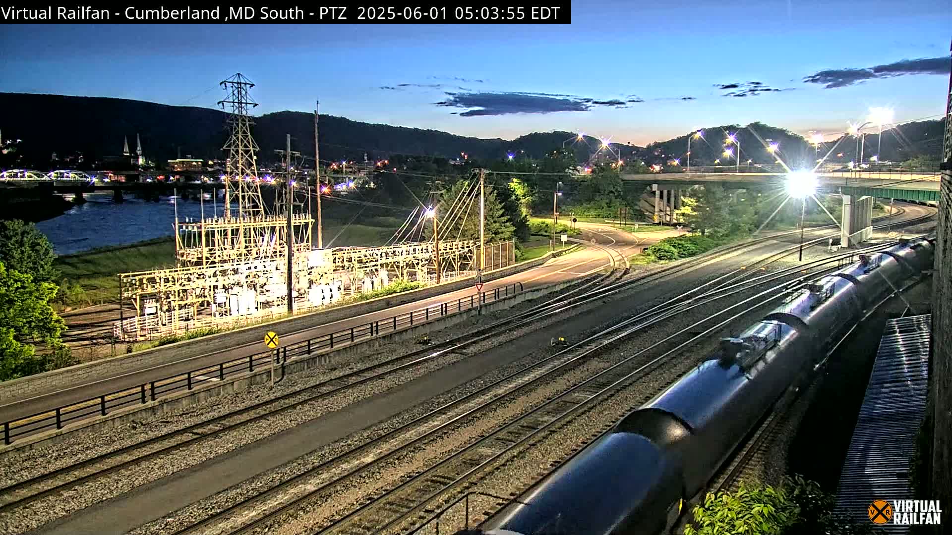 A long train of tanker cars sits on railroad tracks next to an electrical substation at twilight, with a river and a town visible in the background.