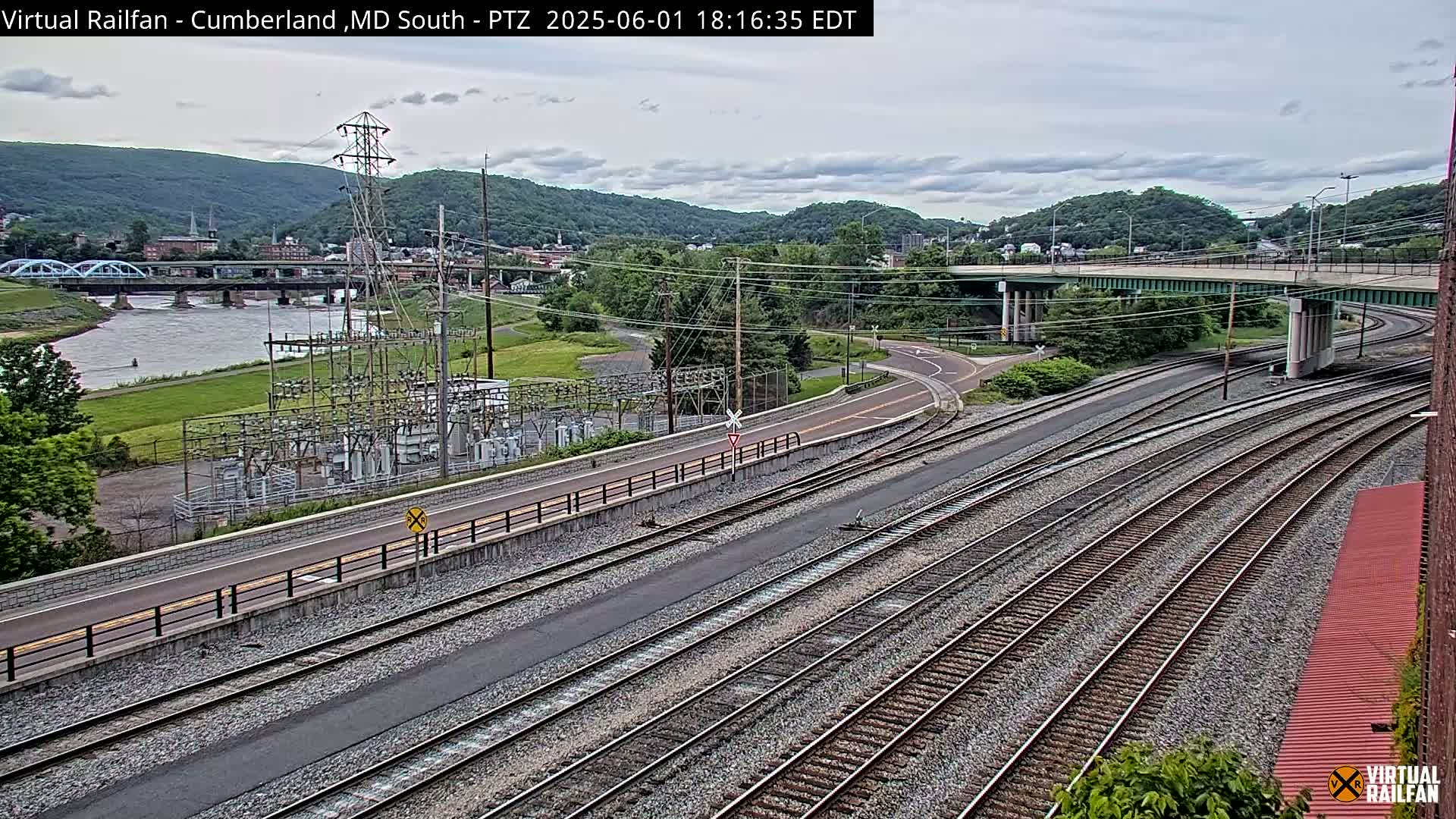 A high-angle view shows multiple sets of train tracks curving around a road and under a bridge, with a river, bridge, and distant hills under a cloudy sky.