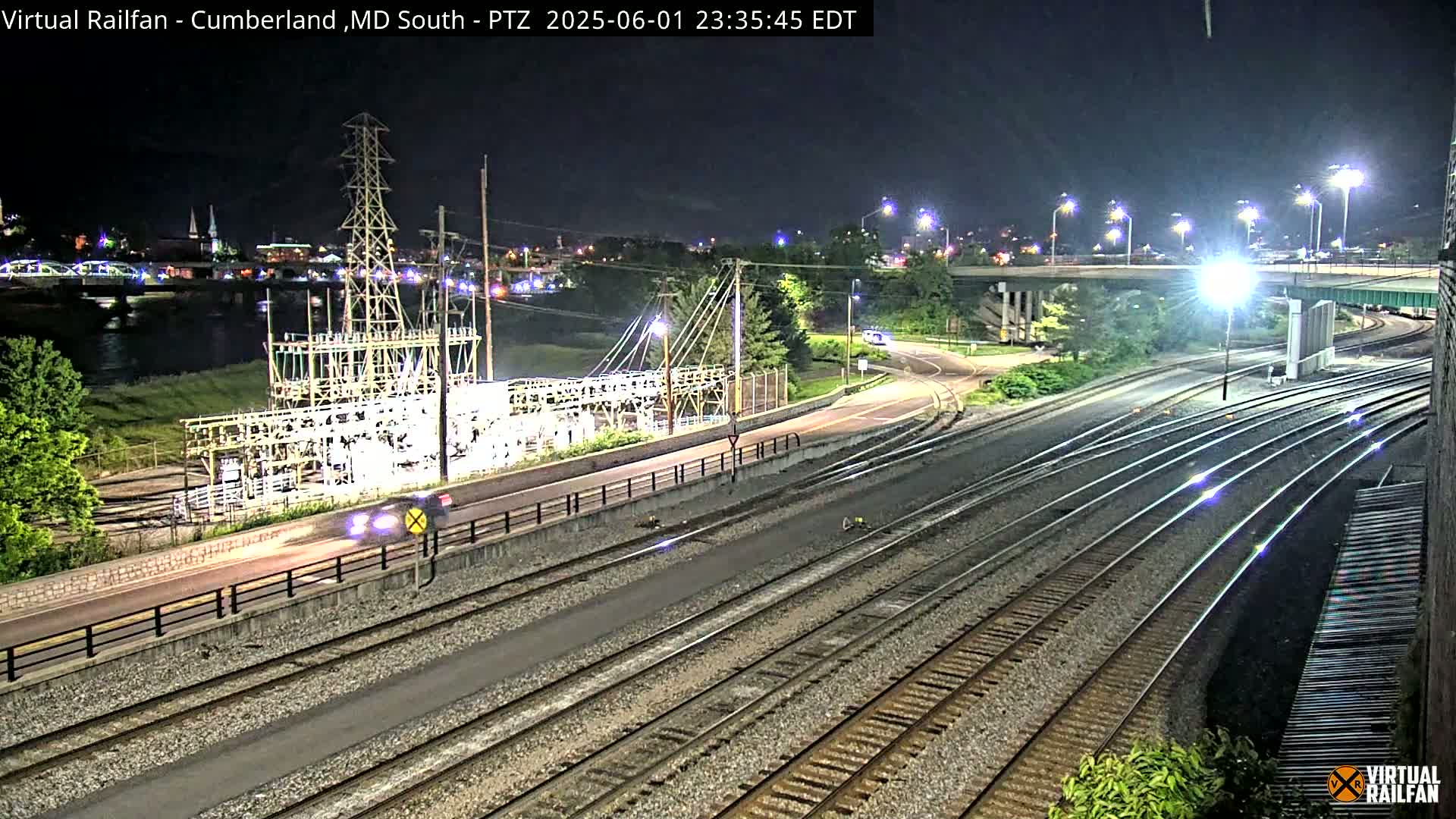 A nighttime view of a rail yard and roadway near a river, with a power substation visible, under clear skies.