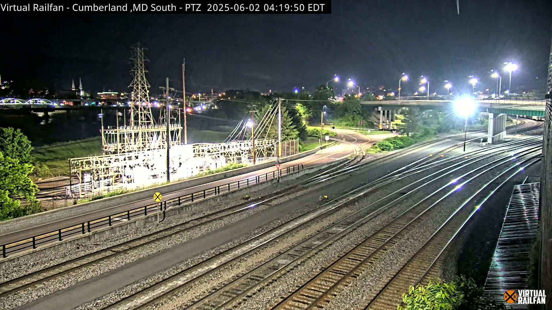 A nighttime view of train tracks alongside a road, passing near an electrical substation and a river with a bridge in the background.