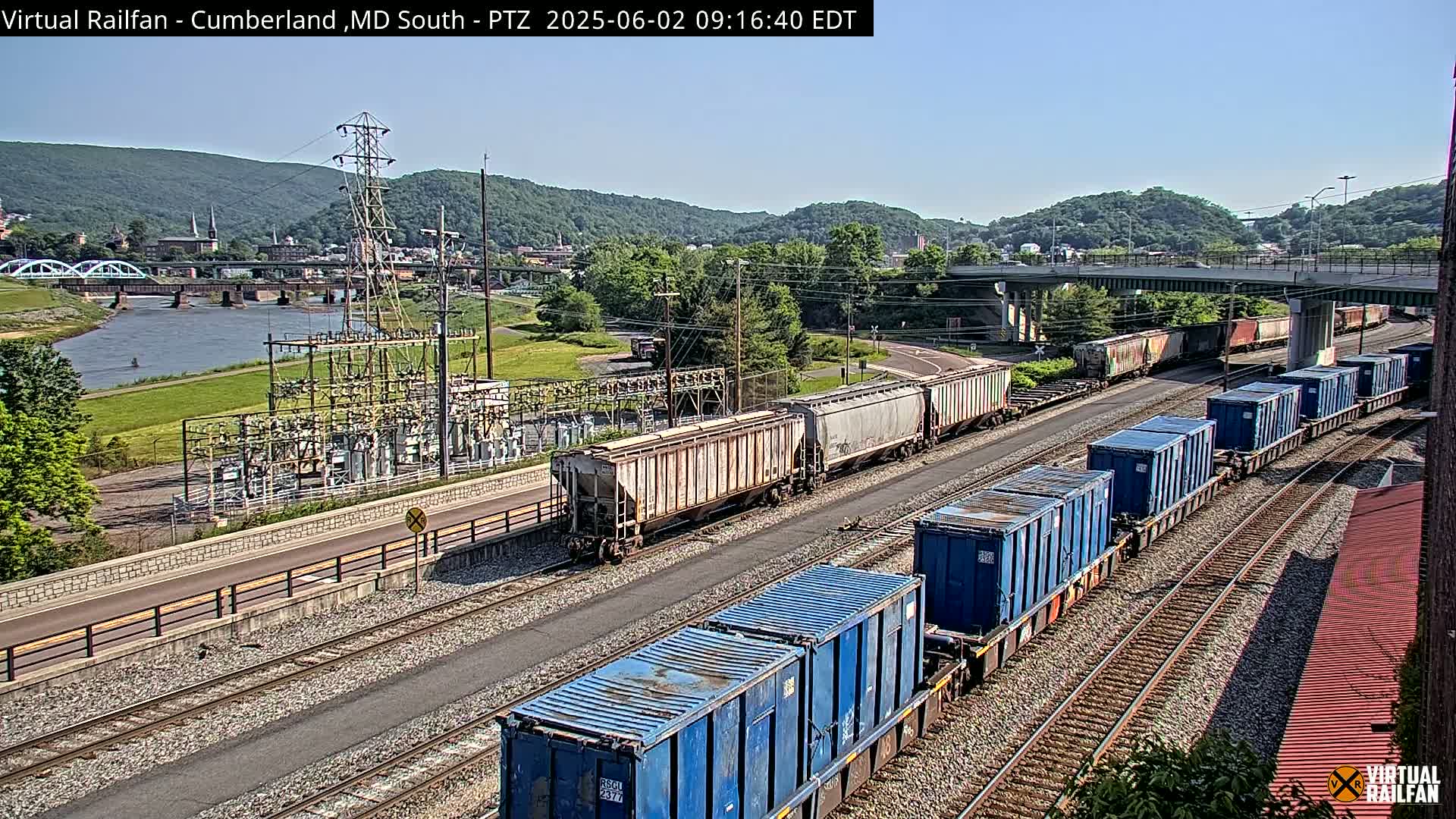 Two freight trains are visible on parallel tracks, near a river and power substation, under a clear sunny sky.