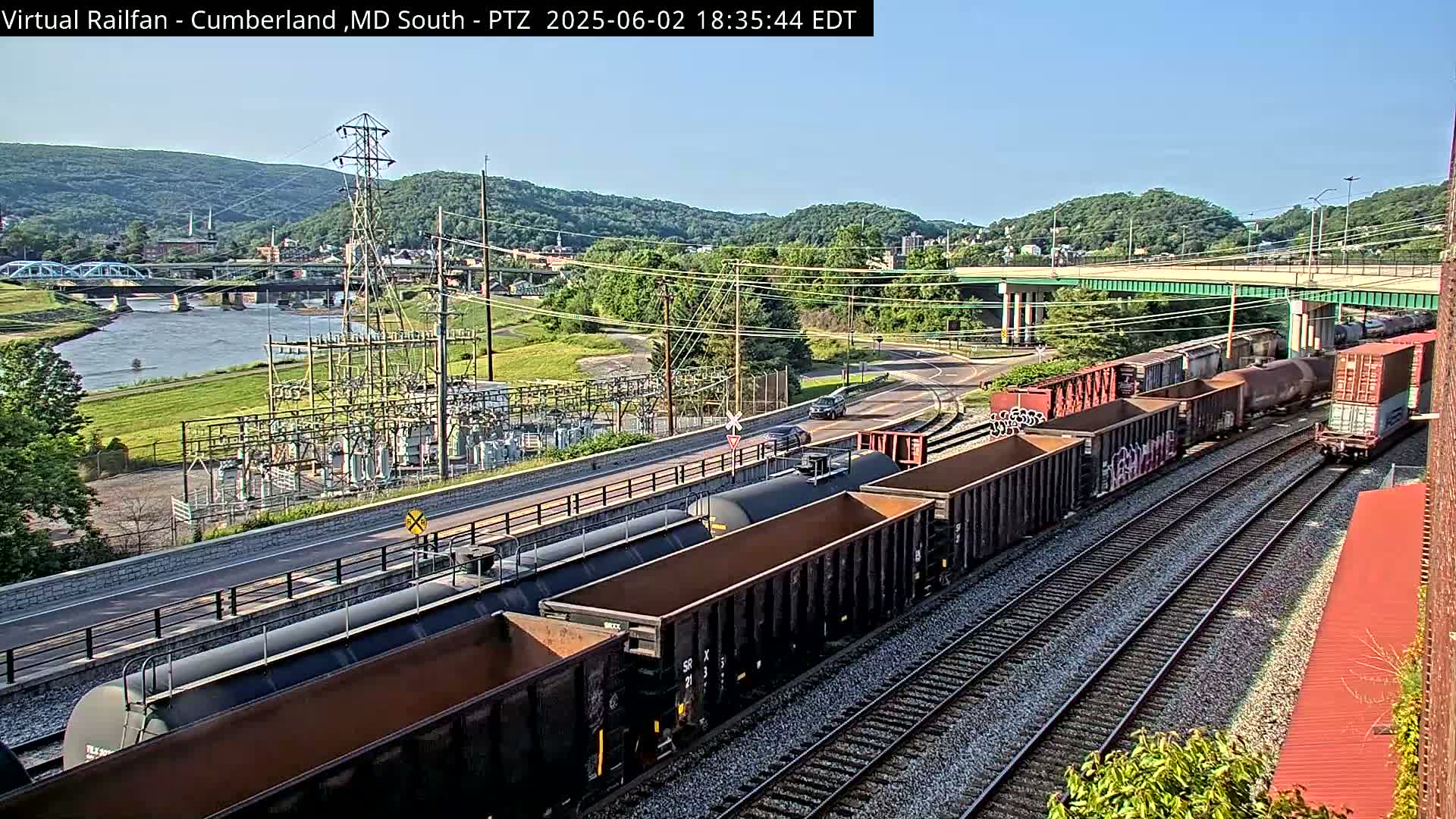 A freight train carrying various cargo cars passes alongside a river and power station under a sunny sky.