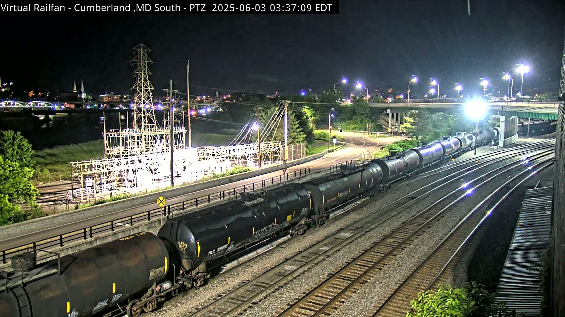 At night, a long freight train of tanker cars travels along railroad tracks near a highway and an electrical substation, with a river and a town visible in the distance.