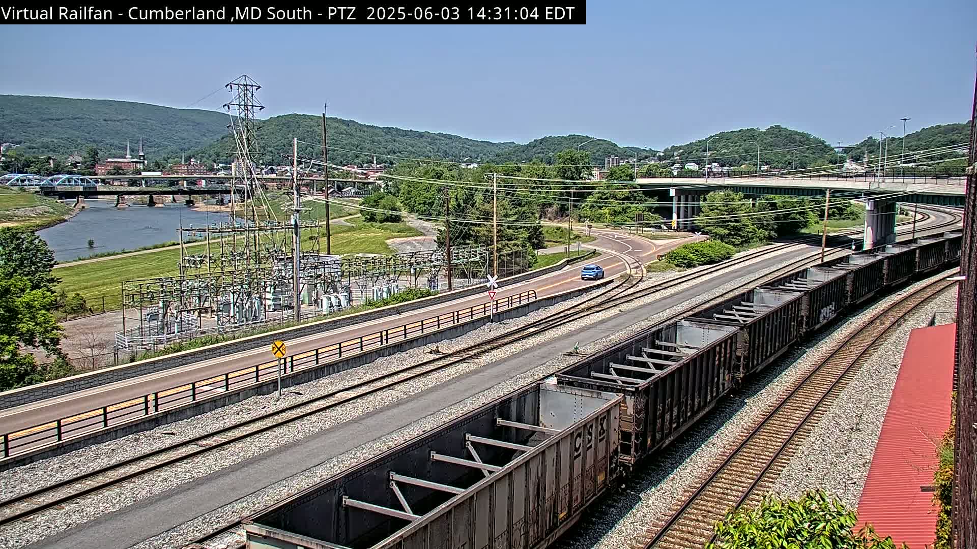 A long train of dark gray freight cars sits on railroad tracks next to a road and river under a clear, sunny sky, with a power substation and a town visible in the distance.