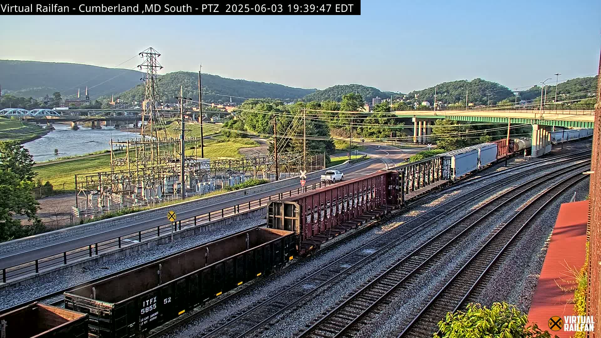 A long freight train sits on multiple parallel train tracks next to a river under a clear, daytime sky, with a highway, power station, and hills in the background.