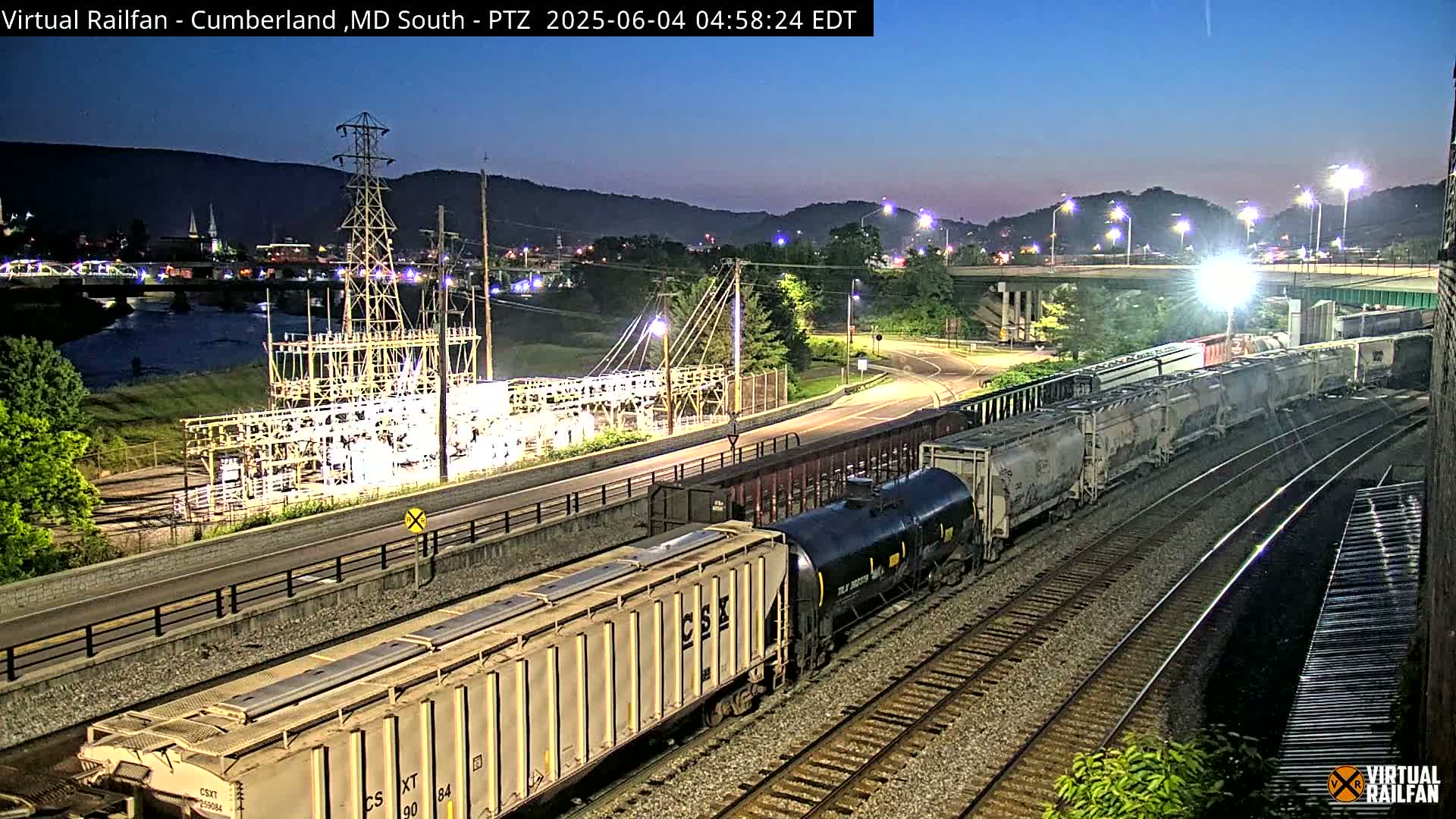 A long freight train is traveling along railroad tracks at night under a twilight sky, passing by a river, a power station, and a highway.