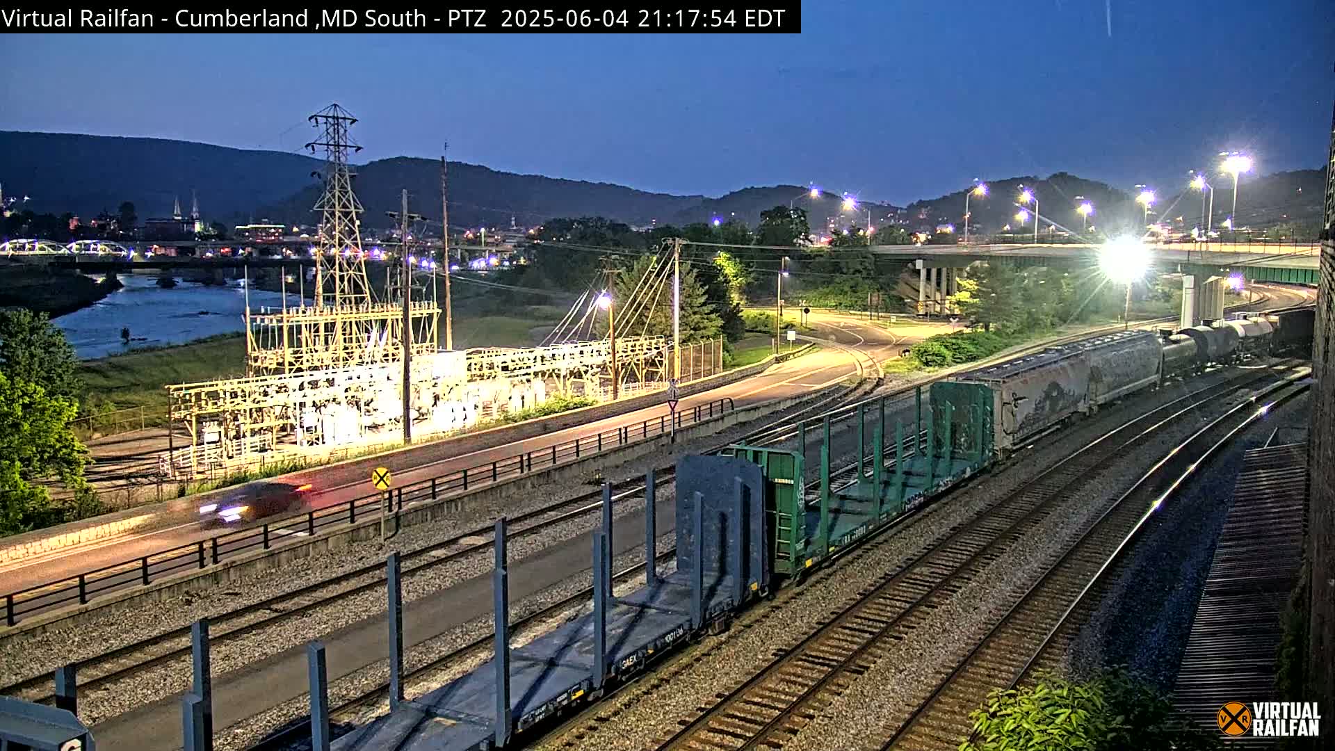 At twilight, a train passes an electrical substation near a river under a highway overpass, with hills in the background.