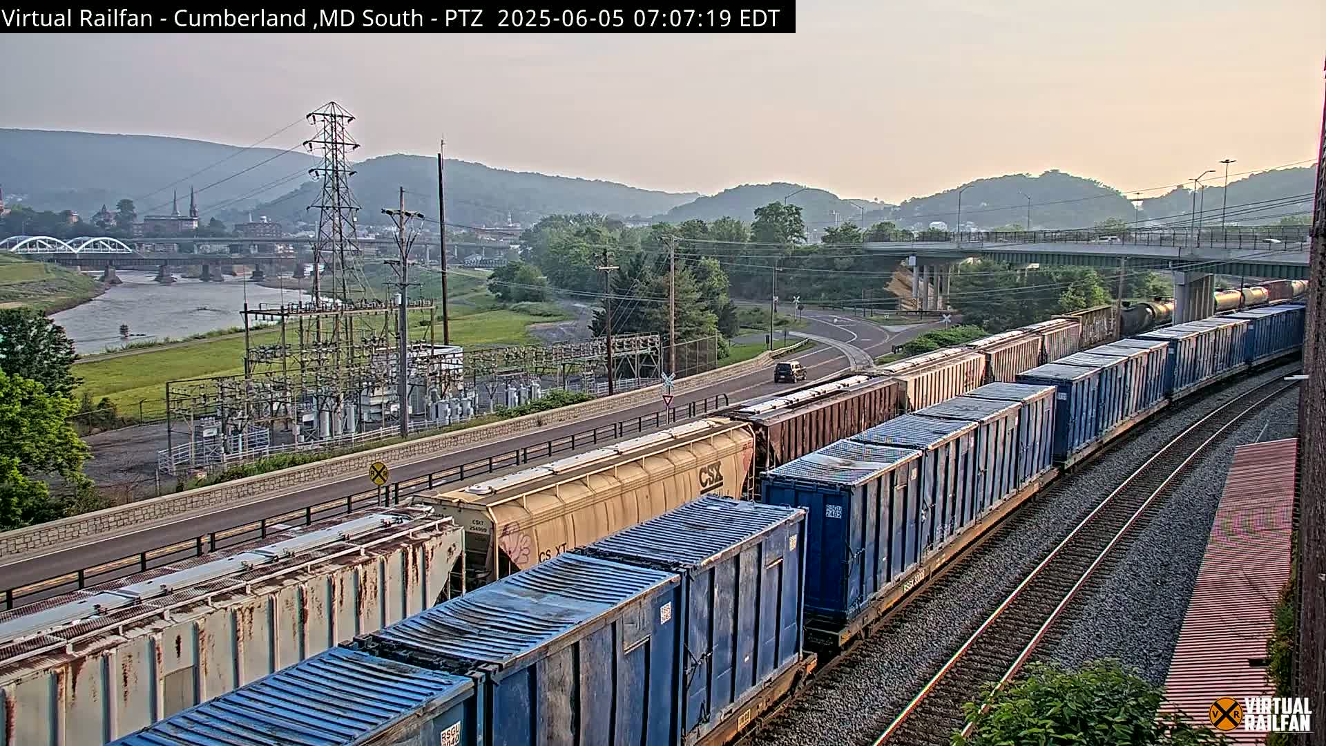 A long freight train carrying blue and tan containers travels along a railway track beside a river, under a pale sky, with a town and hills in the background.